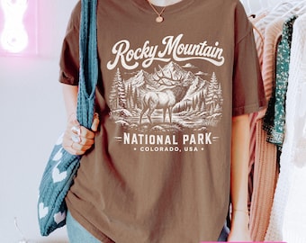 Puede incluir: Una camiseta marrón de manga corta con un estampado gráfico blanco de un alce frente a montañas y pinos. El texto dice Rocky Mountain NATIONAL PARK COLORADO, USA. La camiseta tiene cuello redondo y un corte relajado.