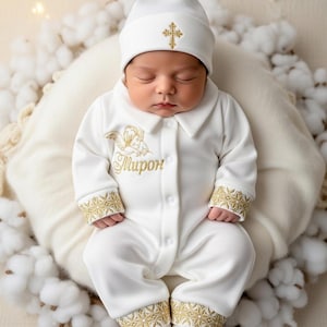 May include: A baby in a white baptismal outfit, featuring a hat and a onesie, decorated with gold crosses and embroidery. The baby is resting on a white pillow surrounded by cotton.
