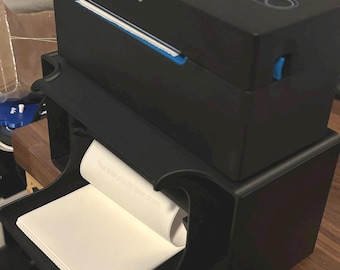 May include: A black label printer with a blue trim and a white paper roll. The printer has a label that says "NELKO" on it. The printer is sitting on a wooden surface.