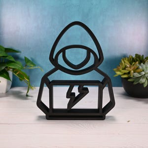 May include: A black, geometric sculpture of a person wearing a hoodie, with a lightning bolt design on the chest. The artwork is displayed on a white surface with a blue background and greenery.