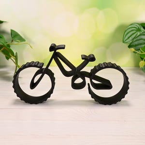 Hollow geometric bicycle sculpture with thick ridged wheels, standing upright on a light wood surface. Set against a soft green bokeh background with leafy plants on both sides. Viewed straight on from the side, showing smooth contours.