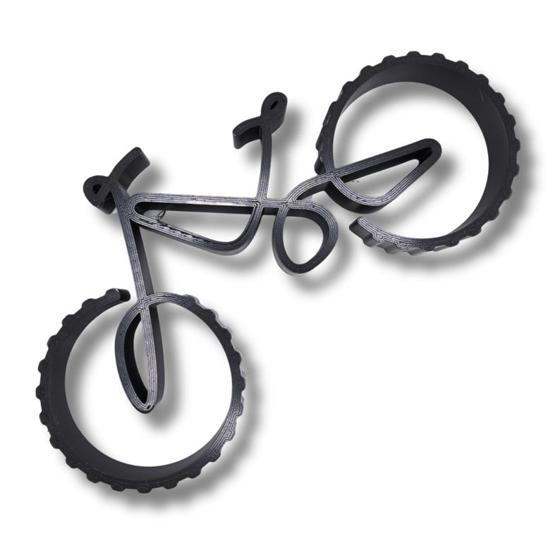 Minimalist bicycle sculpture with hollow frame and thick, ridged wheels, viewed from above at an angle. Displayed on a white surface, showing detailed layering texture and shadows beneath the object.