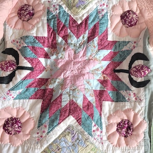 Könnte beinhalten: Handgefertigte Steppdecke mit zentralem Sterndesign in Rosa und Türkis, umgeben von floralen Applikationen. Die Steppdecke weist verschiedene gemusterte Stoffe und einen gesteppten rosa Rand auf und zeigt eine Vintage-Ästhetik.