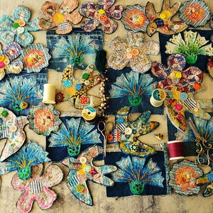 Op de afbeelding: Een verzameling handgemaakte stoffen bloemen en decoratieve elementen, waaronder een schaar, klosjes garen en denim patches. De bloemen hebben verschillende kleuren en patronen, met knoopaccenten. Het algemene thema is knutselen en textielkunst.