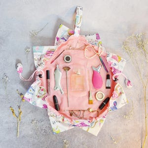 May include: A pink cosmetic bag with a floral and cartoon print, open to display various beauty products. Items include lipstick, perfume, brushes, and a skincare device. The bag is on a gray surface.