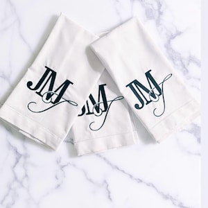 May include: Three white hand towels with the embroidered initials "JM" in a dark teal color. The towels are arranged on a white marble surface. The towels are folded and have a simple, elegant design.