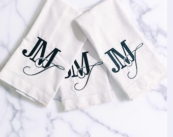 May include: Three white hand towels with the embroidered initials "JM" in a dark teal color. The towels are arranged on a white marble surface. The towels are folded and have a simple, elegant design.