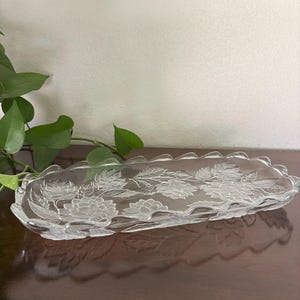 Puede incluir: Bandeja de servir de vidrio transparente con diseño floral. La bandeja rectangular tiene un borde festoneado y está decorada con flores y hojas grabadas. La bandeja se muestra sobre una superficie marrón oscuro.
