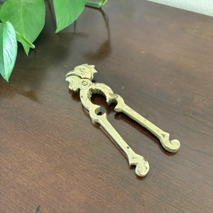 May include: A vintage brass nutcracker with a decorative design. The nutcracker is open, revealing the cracking mechanism. The handles have a curved shape, and the top features a detailed, ornate design. The nutcracker is resting on a dark brown wooden surface.