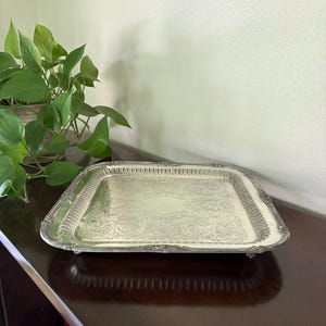 Puede incluir: Una bandeja rectangular plateada con detalles florales ornamentados. La bandeja tiene un borde elevado y se apoya sobre pequeños pies. El interior de la bandeja presenta un patrón repetitivo. La bandeja está sobre una superficie de madera oscura.