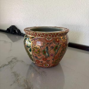 May include: A small, round ceramic bowl featuring a detailed, multi-coloured pattern of butterflies, flowers, and geometric shapes on a salmon-pink background. The rim is decorated with a gold-trimmed, repeating lattice design in red and white.
