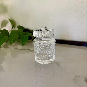 May include: A clear, cut-glass jar with a decorative lid. The jar has a cylindrical body with intricate geometric patterns and a starburst design on the base. The lid features a ringed design and a small, faceted knob. The jar is likely for storing small items.