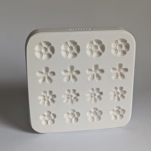 以下が含まれることがあります： 様々な花の模様が施された白い長方形の型。この型には、それぞれ詳細な花びらの模様を持つ12種類の異なる花の形があります。花は3列4行に配置され、対称的で整然とした表示になっています。