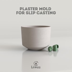以下が含まれることがあります： 「PLASTER MOLD FOR SLIP CASTING」のテキストが入った、スリップキャスティング用の薄いベージュ色の石膏型。型はわずかに傾斜したベースを持つ円筒形です。2つの小さな緑色のプラスチック部品が型の隣にあります。