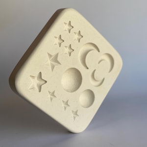 May include: Off-white, diamond-shaped mould featuring celestial designs. The mould has embossed stars, crescent moons, and circular shapes. The design is symmetrical, with the celestial bodies evenly spaced. The mould is made of a textured material.