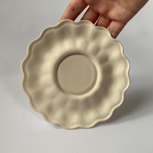 May include: A beige ceramic saucer with a scalloped edge. The center of the saucer has a slightly raised, circular area. The saucer is held by a hand against a white background.