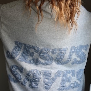 May include: A grey hooded sweatshirt with the words "JESUS SAVES" in blue fabric letters with a floral pattern.