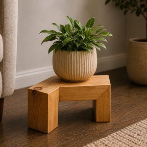 May include: A small, square wooden plant stand with a potted plant. The stand is made of light-colored wood and has a simple, modern design. The plant pot is beige and has vertical grooves. The plant has green leaves.
