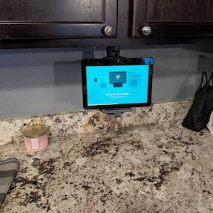 Op de afbeelding: Een tablet aan de muur gemonteerd, met een blauw scherm met een Wi-Fi-symbool en de tekst "Getting Your Device Ready...". De tablet wordt vastgehouden door een zwarte houder die aan een donkere kast is bevestigd. Een roze kaars staat op het aanrecht.