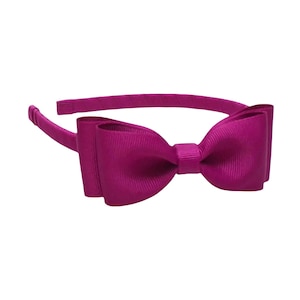 May include: A magenta-colored headband with a large bow. The bow is made of a ribbed fabric and is centered on the headband. The headband is a solid magenta color and is designed to be worn on the head.