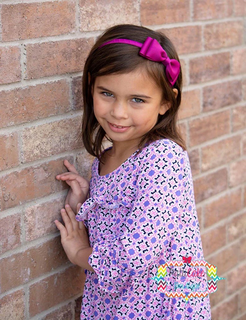 Puede incluir: Una joven con una diadema morada con lazo y un vestido morado de manga larga con estampado floral. El fondo es una pared de ladrillos rojos.