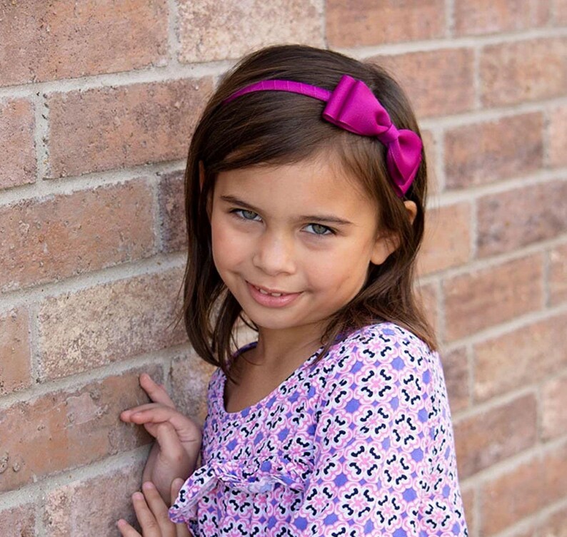 Puede incluir: Una ni&ntilde;a con cabello casta&ntilde;o y ojos azules sonr&iacute;e, usando una diadema con lazo magenta. Su vestido morado y blanco con estampado complementa la diadema. El fondo presenta una pared de ladrillos.