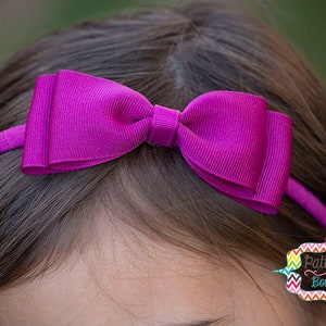 Puede incluir: Una diadema magenta con un lazo grande. El lazo y la diadema est&aacute;n hechos de tela acanalada. La diadema se lleva en la cabeza de una persona. El logotipo de PattiCake Boutique es visible en el lado derecho de la imagen.