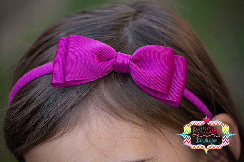 Puede incluir: Una diadema magenta con un lazo grande. El lazo y la diadema est&aacute;n hechos de tela acanalada. La diadema se lleva en la cabeza de una persona. El logotipo de PattiCake Boutique es visible en el lado derecho de la imagen.