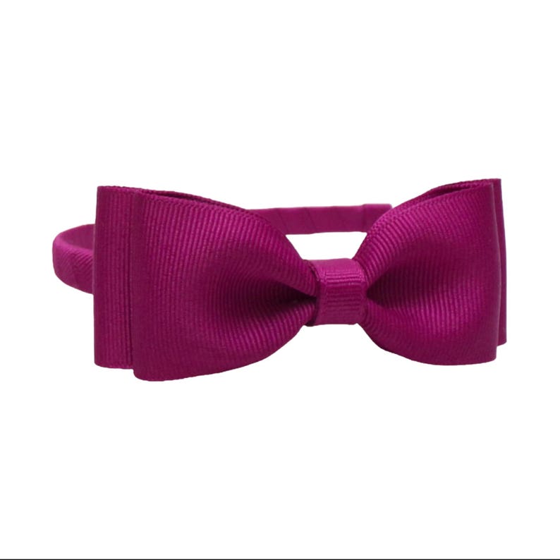 Puede incluir: Una pajarita magenta con textura acanalada, sujeta a una diadema a juego. La pajarita es sim&eacute;trica y est&aacute; centrada. El accesorio es de un vibrante tono magenta.