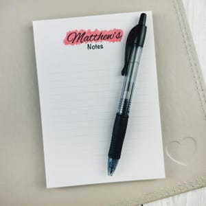 May include: A white rectangular notepad with horizontal lines and a pink watercolour brushstroke graphic featuring the black text Matthew's Notes. A black retractable ballpoint pen with a clear barrel and a black rubber grip rests on the notepad.