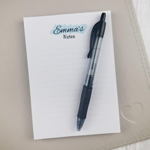 May include: A white rectangular notepad with horizontal lines and a blue watercolor-style accent behind the text Emma's Notes. A black retractable ballpoint pen with a clear barrel and a black rubber grip is placed diagonally across the notepad.