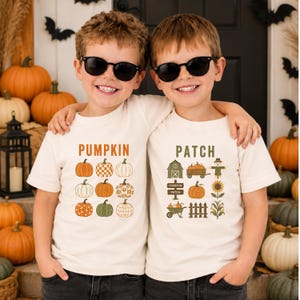 May include: Two off-white short-sleeve t-shirts featuring autumn-themed graphics. The left shirt displays the word PUMPKIN above a grid of nine pumpkins in various patterns. The right shirt displays the word PATCH above icons of a barn, wagon, scarecrow, sign, pumpkin, sunflower, wheelbarrow, fence, and corn.