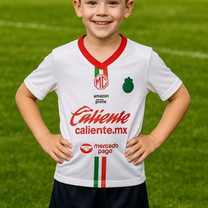 Może przedstawiać: Dziecko w białej koszulce piłkarskiej z czerwonymi i zielonymi akcentami oraz napisem "Caliente caliente.mx". Koszulka ma zielone logo i pasujące logo na granatowych spodenkach. Tło to zielone pole.