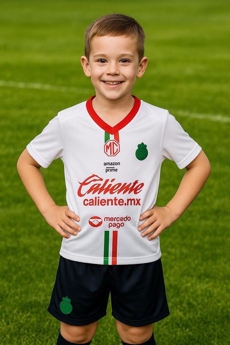 Może przedstawiać: Dziecko w białej koszulce piłkarskiej z czerwonymi i zielonymi akcentami oraz napisem "Caliente caliente.mx". Koszulka ma zielone logo i pasujące logo na granatowych spodenkach. Tło to zielone pole.