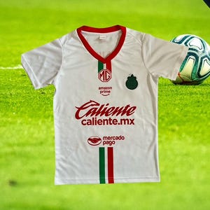 Może przedstawiać: Biała koszulka piłkarska z czerwonymi i zielonymi akcentami. Koszulka zawiera napis "Caliente caliente.mx" w czerwonym skrypcie, logo Amazon Prime i emblemat drużyny. Piłka nożna jest widoczna w tle.