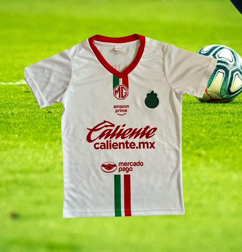 Może przedstawiać: Biała koszulka piłkarska z czerwonymi i zielonymi akcentami. Koszulka zawiera napis "Caliente caliente.mx" w czerwonym skrypcie, logo Amazon Prime i emblemat drużyny. Piłka nożna jest widoczna w tle.