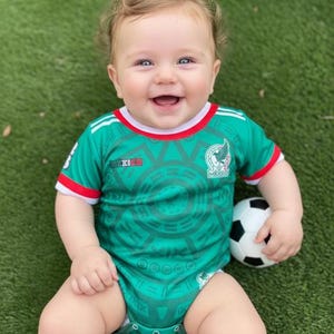 May include: A smiling baby wearing a green and white Mexico soccer jersey, holding a small soccer ball. The jersey has the Mexican national team logo and the word "MEXICO" on it. The baby is sitting on green grass.