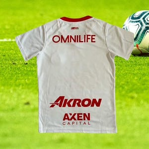 Może przedstawiać: Biała koszulka piłkarska z czerwonymi wykończeniami i napisami. Tył koszulki przedstawia słowo "OMNILIFE" w czerwonych literach blokowych u g&oacute;ry, z "AKRON" i "AXEN CAPITAL" w kolorze czerwonym poniżej. Piłka nożna jest widoczna po prawej stronie.