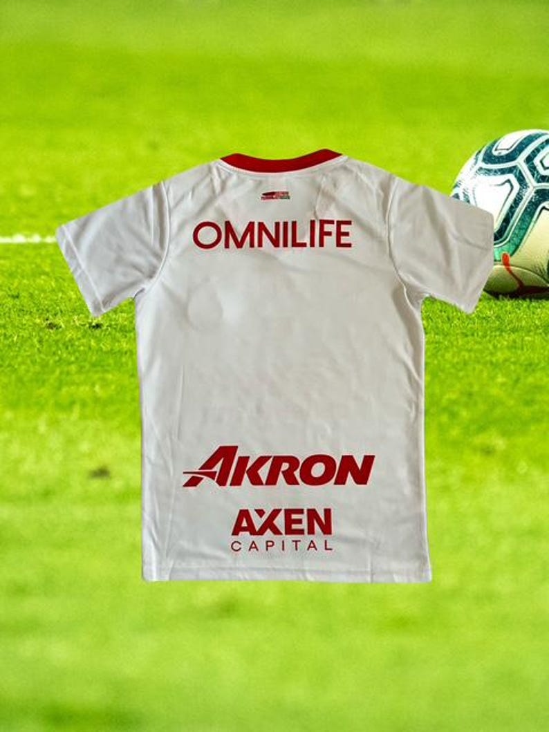 Może przedstawiać: Biała koszulka piłkarska z czerwonymi wykończeniami i napisami. Tył koszulki przedstawia słowo "OMNILIFE" w czerwonych literach blokowych u g&oacute;ry, z "AKRON" i "AXEN CAPITAL" w kolorze czerwonym poniżej. Piłka nożna jest widoczna po prawej stronie.