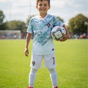 May include: A young person wearing a white and teal soccer uniform with the number 7 on the shorts. The uniform features the Portuguese coat of arms. The person is holding a soccer ball and standing on a green field.