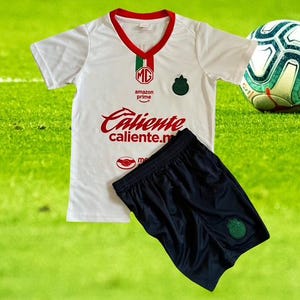 Może przedstawiać: Biała koszulka piłkarska i granatowe szorty z czerwonymi i zielonymi akcentami. Koszulka ma czerwony napis "Caliente" i logo. Piłka nożna znajduje się w tle na zielonym boisku.