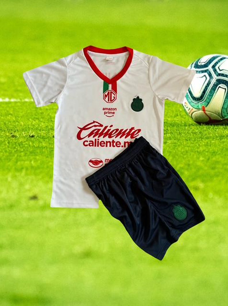 Może przedstawiać: Biała koszulka piłkarska i granatowe szorty z czerwonymi i zielonymi akcentami. Koszulka ma czerwony napis "Caliente" i logo. Piłka nożna znajduje się w tle na zielonym boisku.