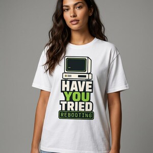 Peut inclure: Un t-shirt blanc à manches courtes arborant un graphique représentant un écran d'ordinateur vintage au-dessus du texte "HAVE YOU TRIED REBOOTING" en lettres noires et vertes. Le motif est centré sur la poitrine, porté avec un jean bleu.