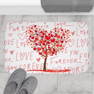 May include: A white bath mat with a tree design made of red hearts. The words "Love," "Forever," and "Adore" are written in red script across the mat. Gray slippers are at the bottom.