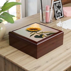 May include: A wooden jewelry box with a glossy, dark brown finish. The lid features a vibrant painting of a sunflower with yellow petals and green leaves. The box is rectangular with rounded edges, sitting on a wooden surface.