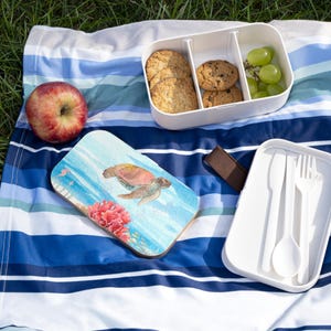 May include: A picnic setting featuring a white lunchbox with cookies and grapes, a wooden board with a sea turtle design, a red apple, and a white cutlery container. These items are arranged on a blue and white striped blanket.