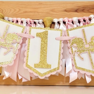 May include: A pink and gold glitter banner with the number "1" and two carousel horses. The banner is decorated with pink and white satin ribbon streamers.