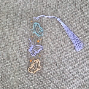 May include: A clear rectangular bookmark featuring three butterfly illustrations in light blue, lavender, and peach colors, accented with small orange diamond shapes and stars, finished with a lavender tassel attached to the top.