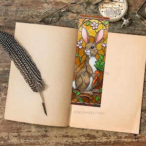 May include: A stained-glass style bookmark featuring a brown bunny holding a carrot. The bookmark has pink flowers and green leaves. The bookmark is placed in an open book with a feather quill and glasses nearby. The text "@IRONWOOD INK" is visible.