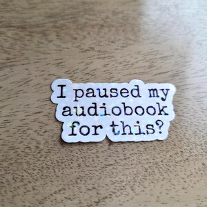 Pu&ograve; includere: Un adesivo fustellato con finitura olografica che riporta il testo nero "I paused my audiobook for this?". L'adesivo presenta un bordo bianco e un sottile motivo a stelle integrato nelle lettere su uno sfondo scintillante e riflettente.